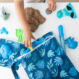 A child plays with kinetic sand and blue beach toys near a Bumkins Wet Bag: Blue Tropic on a white table.