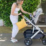 A woman in white hands an item to a baby in a stroller with a Bumkins Wet Bag: Sunshine on the handle outdoors.