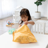 A young girl at a white table opens a Bumkins Wet Bag: Sunshine, with a playhouse and toys brightening the airy room.