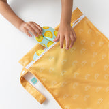 Close-up of a child's hands tucking a colorful sandal into a Bumkins Wet Bag: Sunshine on a white table.