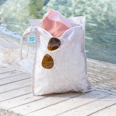A bag from Bumkins Wet Bag 2 Pack: Wander and Sunshine holds sunglasses and swimwear poolside with empty lounge chairs in the background.