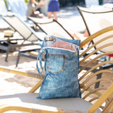 A bag from Roamio by Bumkins Wet Bag + Wet/Dry Clutch Set: Denim On Denim holds a water bottle on a lounge chair poolside.