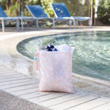 A bag from Bumkins The Chaos Coordinator Essential 4-Piece Set: Lace holds a water bottle, swimsuit, & towel poolside; lounge chairs behind.