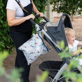 A person in black overalls pushes a stroller with a toddler while a Bumkins Wet Bag: No Tricks, Just Treats hangs from the handle.