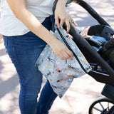 Wearing jeans and a white shirt, a person unzips a Bumkins Wet Bag: No Tricks, Just Treats attached to a stroller outdoors.