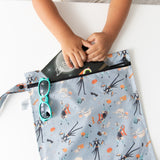 A child slips a book into a Bumkins Wet Bag: No Tricks, Just Treats on a white table, with blue sunglasses nearby.