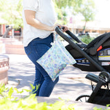A person pushes a stroller outdoors with a Bumkins Disney Aloha Stitch Wet Bag + Clear Travel Bag 3 Pack Set on the handle.