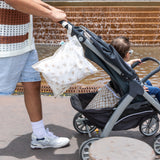 An adult pushes a stroller with a baby near a fountain, with Bumkins Wet Bag 2 Pack: Palm Check hanging from the handle and placed underneath.