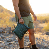 Person outdoors with a Roamio by Bumkins Wet Bag 2 Pack: Desert Night, wearing green shorts and black shirt in sunlight.