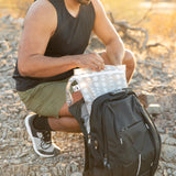 Outdoors, someone packs a Roamio by Bumkins Wet Bag 2 Pack: Desert Night into a black backpack on rocky ground.