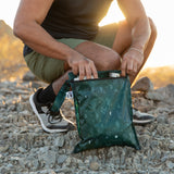 Kneeling outdoors, someone opens a Roamio by Bumkins Wet Bag 2 Pack: Desert Night, made from waterproof fabric.
