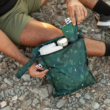 Person sitting on rocky ground, holding a green patterned bag with a water bottle and towel partially visible inside.