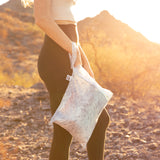 A woman in athletic wear holds a Roamio by Bumkins Wet Bag 2 Pack: Disco Boots on rocky terrain in golden sunlight.