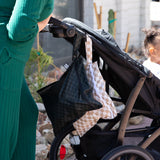 An adult strolls outdoors with Bumkins Wet Bag 2 Pack in Sand & Black Check hanging on the stroller; child rides comfortably.