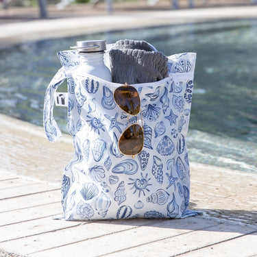 A beach bag with blue seashell patterns holds a gray towel, water bottle, and sunglasses by a poolside.