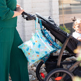 Woman in green pushes stroller with baby and Bumkins Wet Bag 2 Pack: Ocean Life and Jungle hanging from handle on a sunny day.