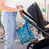 A parent pushes a stroller with a Bumkins Wet Bag 2 Pack: Ocean Life and Jungle, featuring waterproof fabric, hanging on the handle.