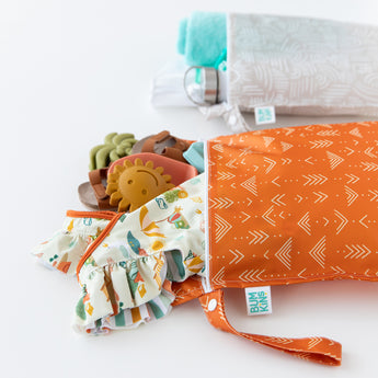 Bumkins Wet Bag 2 Pack: Wander + Grounded sits on a white background, with the orange bag in the foreground holding toys and clothes.