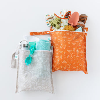 Bumkins Wet Bag 2 Pack: Wander + Grounded, filled with beach essentials like goggles, a towel, clothes, and toys, lay on a white background.