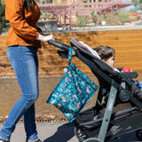 A woman pushes a stroller with a baby and a Bumkins Wet Bag 2 Pack: Jungle + Animal Prints hanging from the handle by an outdoor water feature.