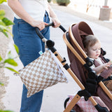 A person pushes a toddler in a stroller outdoors with a Bumkins Wet Bag Plus 2 Pack: Mauve + Sand Check hanging on the handle.