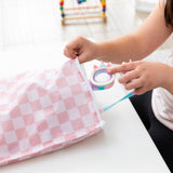 A child places goggles into a Bumkins Wet Bag Plus 2 Pack: Mauve + Sand Check on a white table.