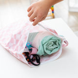 A child's hand holds a Bumkins Wet Bag Plus 2 Pack: Mauve + Sand Check open on a white table, revealing a swimsuit and towel inside.