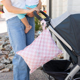 A woman holds a baby outdoors by a stroller with a Bumkins Wet Bag Plus 2 Pack: Mauve + Sand Check hanging from the handle.