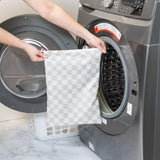 A person holds a Bumkins Wet Bag Plus 2 Pack: Mauve + Sand Check in front of an open front-loading washing machine.
