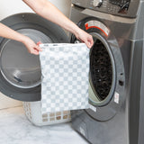 A person holds a Bumkins Wet Bag Plus 2 Pack: Mint + Charcoal Check in front of an open front-loading washing machine.