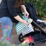 A woman places a stuffed toy into Bumkins Wet Bag Plus 2 Pack: Green + Gray Stripe attached to a stroller with a baby outdoors.