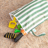 A Bumkins Wet Bag Plus 2 Pack: Green + Gray Stripe lays open on sand with beach toys spilling out.