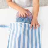 A child places glittery sandals into a Bumkins Wet Bag Plus 2 Pack: Blue + Yellow Stripe on a white table.