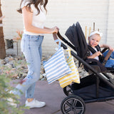 A woman pushes a stroller outdoors with a smiling toddler and Bumkins Wet Bag Plus 2 Pack: Blue + Yellow Stripe hanging on the handle.