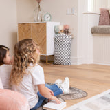 Children sit on a rug with books, facing a cabinet with Bumkins Extra Large Wet Bag: Charcoal Check holding stuffed animals nearby.