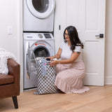 In a cozy laundry room, a person kneels to load clothes into a front-loading washer from a Bumkins Extra Large Wet Bag: Charcoal Check.