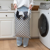 In a laundry room, a person holds a Bumkins Extra Large Wet Bag: Charcoal Check filled with clothes near a washing machine.