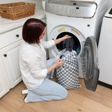 A woman kneels by a front-loading washer, placing clothes from a Bumkins Extra Large Wet Bag: Charcoal Check inside.