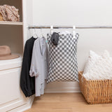 A closet corner with shirts and a Bumkins Extra Large Wet Bag: Charcoal Check hanging on a rod, a basket with pillows, and a hat on shelf.