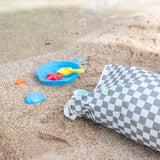 A beach scene with a Bumkins Extra Large Wet Bag: Charcoal Check, a blue bucket, yellow shovel, and colorful toys on sand by the water's edge.