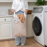 A person with a Bumkins Extra Large Wet Bag: Wander, featuring adjustable handles, stands by a washing machine in a laundry room.