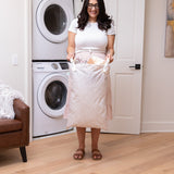 A person smiling, holding a Bumkins Extra Large Wet Bag: Wander filled with clothes in front of a washer and dryer.