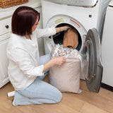 A person kneels by a front-loading washer, placing clothes inside from a Bumkins Extra Large Wet Bag: Wander, made of waterproof fabric.