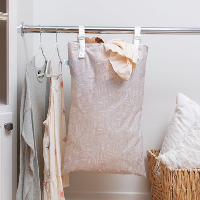 A beige patterned laundry bag hangs from a clothing rack with white straps. Next to it, shirts and dresses hang on hangers. A wicker basket is on the floor nearby. The background is a plain light-colored wall.