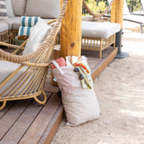 Bumkins Extra Large Wet Bag: Wander, filled with beach essentials, sits by wicker seating on a wooden deck framed by sand and rope.