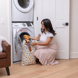 A woman in a white shirt and pink skirt uses a Bumkins Extra Large Wet Bag: Winnie + Friends to load laundry in a modern washroom.