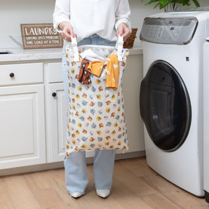 A person holding a Bumkins Extra Large Wet Bag: Winnie + Friends with clothes in a modern laundry room, between a washing machine and cabinet.