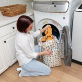 A person kneels by a front-load washer, placing colorful clothes into a Bumkins Extra Large Wet Bag: Winnie + Friends.