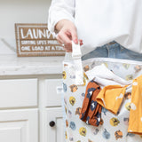 Close-up of a person holding a Bumkins Extra Large Wet Bag: Winnie + Friends filled with clothes in a bright laundry room.