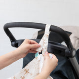 A person attaches Bumkins Wet/Dry Bag: Palm Daze to a stroller handle with the snap closure on the strap.